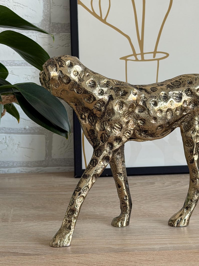 May include: A gold cheetah sculpture with black spots. The decorative figurine is made of metal and has a textured surface. The cheetah is posed in a walking position, with its head turned to the side. The sculpture is a home decor item.