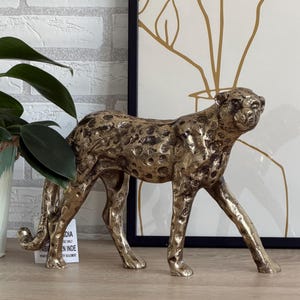 May include: A gold-toned cheetah ornament with dark spots, depicted in a walking pose. The metal sculpture has a textured finish. The cheetah is positioned on a wooden surface, with a plant and artwork visible in the background.
