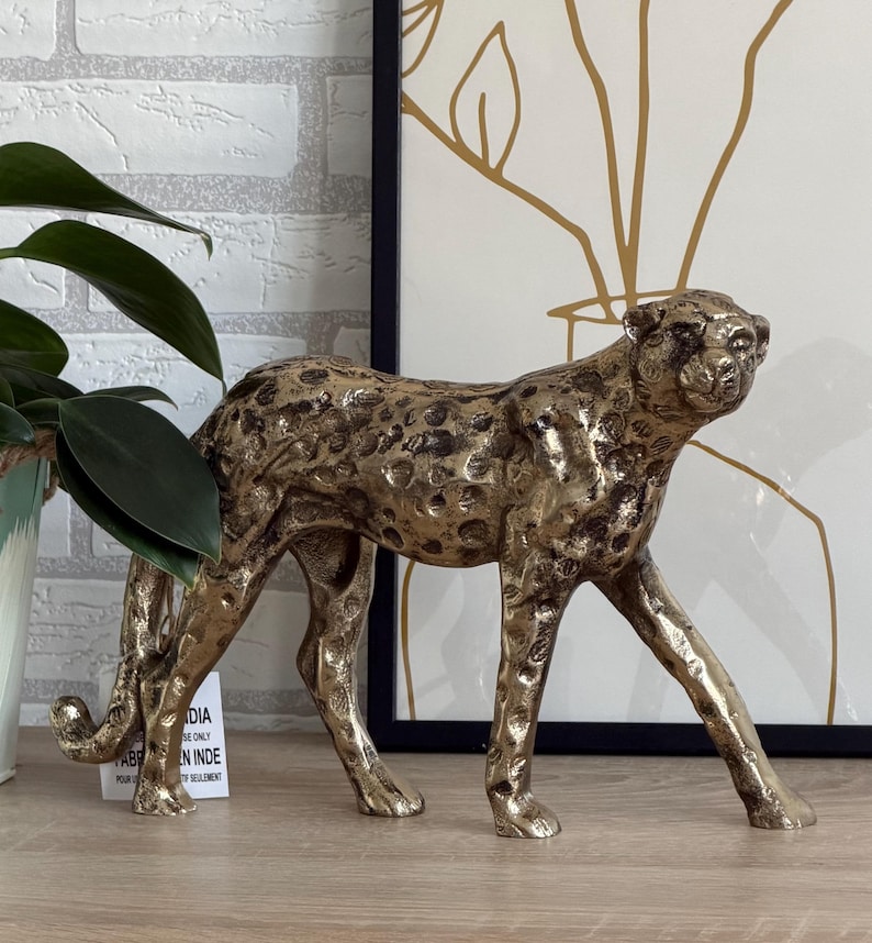 May include: A gold-toned cheetah ornament with dark spots, depicted in a walking pose. The metal sculpture has a textured finish. The cheetah is positioned on a wooden surface, with a plant and artwork visible in the background.