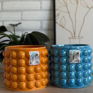 Könnte beinhalten: Zwei Keramik-Blumentöpfe, einer orange und einer blau, mit einem strukturierten, blasenartigen Muster. Beide haben ein kleines Etikett mit der Aufschrift "PORTUGAL". Die Töpfe sind zylindrisch und haben einen Durchmesser von etwa 20 cm.