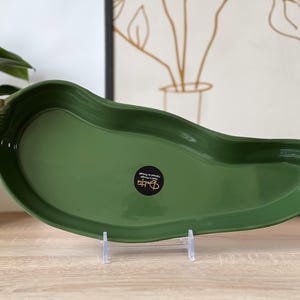 Op de afbeelding: Groene keramische serveerschaal in de vorm van een paprika. De schaal heeft een glanzende afwerking en een bruine en gouden steel. De schaal wordt gepresenteerd op een transparante standaard. Een plant en ingelijste kunst staan op de achtergrond.