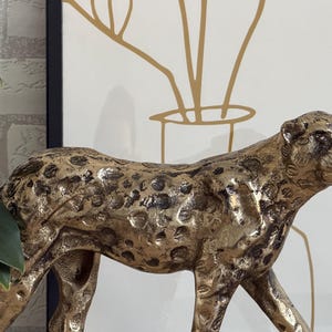 May include: A gold-coloured cheetah figurine with dark spots. The sculpture is detailed and textured, with a focus on the animal's form. The background includes a framed art piece with gold line art and a green plant.