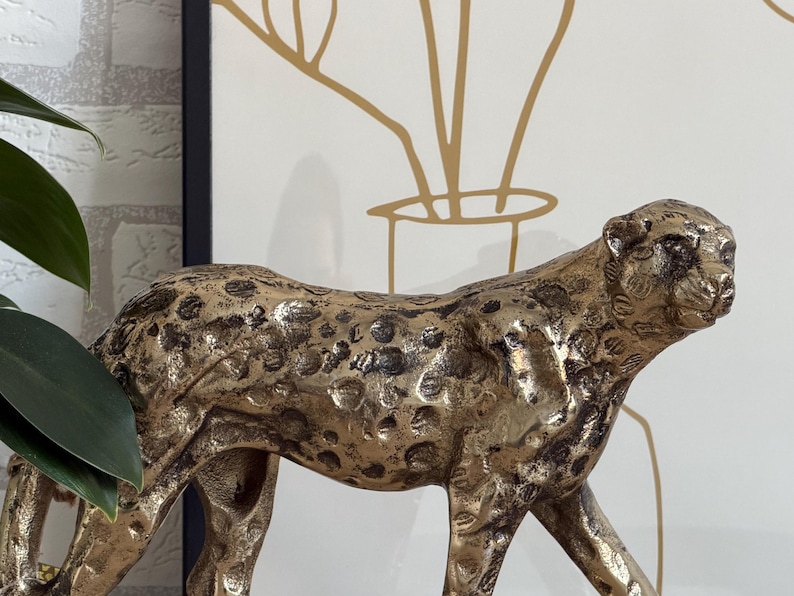 May include: A gold-coloured cheetah figurine with dark spots. The sculpture is detailed and textured, with a focus on the animal's form. The background includes a framed art piece with gold line art and a green plant.