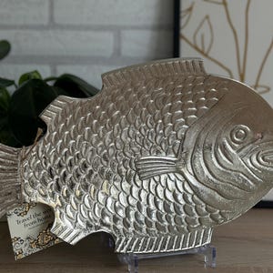 Pode incluir: Uma bandeja decorativa de metal prateado em forma de peixe com escamas, barbatanas e traços faciais em relevo. Uma pequena etiqueta retangular presa à cauda com um cordel apresenta o texto impresso, Travel the world from home.