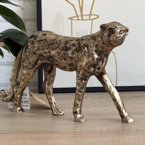 May include: A gold-coloured cheetah figurine with black spots. The sculpture is made of metal and has a textured surface. The cheetah is in a walking pose, with its head turned slightly. A decorative item.