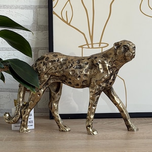 May include: A gold cheetah figurine with black spots, in a walking pose. The sculpture has textured fur and sits on a wooden surface. A framed art print with a gold plant design is in the background.