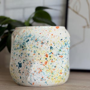 Könnte beinhalten: Eine cremefarbene Keramikvase, verziert mit bunten Farbspritzern in Blau, Orange und Gelb. Die Vase hat eine zylindrische Form mit leicht strukturierter Oberfläche. Ideal zum Halten von Blumen oder als dekoratives Element.