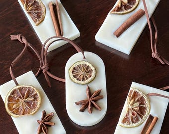 Puede incluir: Cinco velas de cera blancas, rectangulares y ovaladas, decoradas con rodajas de cítricos secos, anís estrellado y palitos de canela. Cada una tiene un cordón de cuero marrón. Dispuestas sobre una superficie de madera oscura.
