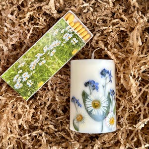 May include: A white pillar candle decorated with pressed flowers, including daisies and blue forget-me-nots. A box of matches with a daisy design is next to the candle, all nestled in brown packing material.
