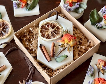May include: White, rectangular wax melts adorned with dried orange slices, red roses, and lavender. Some melts are presented in a brown cardboard box with shredded paper. Brown leather cords are attached for hanging.
