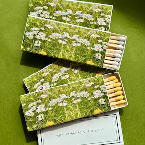 Può includere: Quattro scatole di fiammiferi con un design floreale con margherite bianche in un campo verde. Le scatole di fiammiferi sono aperte, rivelando i fiammiferi con punte gialle. La scatola inferiore ha il testo "my cosy CANDLES" in nero.