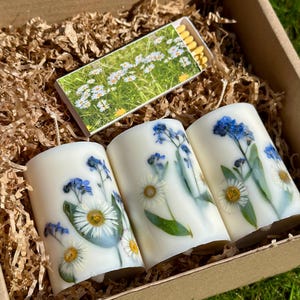 May include: A gift box containing three white pillar candles decorated with pressed daisies and blue forget-me-nots. A box of matches with a floral design is also included. The box is filled with brown paper filler.