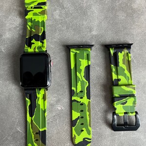 Könnte beinhalten: Eine schwarze Smartwatch mit einem hellgrünen Camouflage-Muster-Armband. Zwei zusätzliche Armbänder mit dem gleichen Muster werden ebenfalls angezeigt. Die Armbänder haben schwarze Schnallen und Anschlüsse.