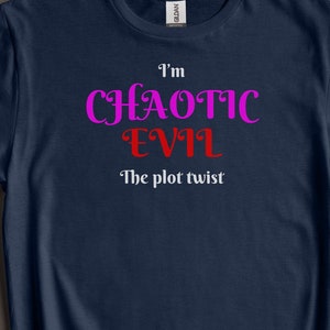 May include: Navy blue t-shirt with the text "I'm Chaotic Evil The plot twist" in pink and red lettering. The shirt is displayed on a neutral background with green foliage.