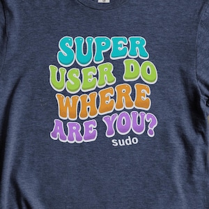 May include: A navy blue t-shirt with the text "SUPER USER DO WHERE ARE YOU?" in colorful bubble letters. The word "sudo" is below the main text. The shirt is surrounded by green leaves and brown stems.