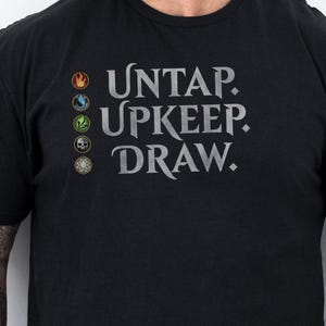 May include: Black t-shirt with the text "UNTAP. UPKEEP. DRAW." in gray. The shirt has four small colored symbols above the text. The shirt is short-sleeved and appears to be made of cotton.