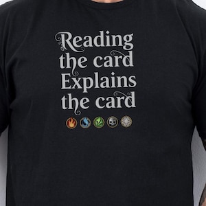 May include: Black t-shirt with the text "Reading the card Explains the card" in a stylized font. Below the text are five small symbols. The shirt is short-sleeved and appears to be made of a soft material.