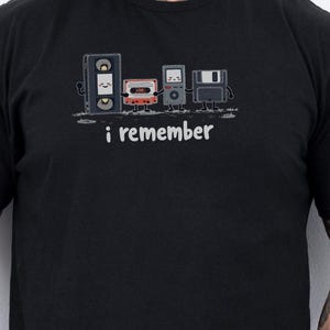 May include: Black t-shirt with a graphic of retro audio equipment, including speakers, a cassette tape, and a floppy disk. The text "i remember" is printed below the graphic in a handwritten style. The shirt is made of a soft material.
