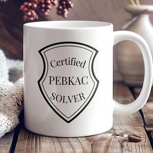 May include: White ceramic coffee mug with a black shield design that says "Certified PEBKAC SOLVER".