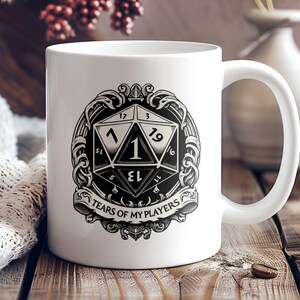May include: White ceramic mug with a black and white graphic of a 20-sided die with the number 1 rolled. The graphic is surrounded by a decorative border and the text "Tears of My Players".