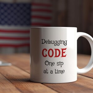 Może przedstawiać: Biały ceramiczny kubek do kawy z czarnym tekstem, kt&oacute;ry brzmi "Debugging CODE One sip at a time". Słowo "CODE" jest czerwone.