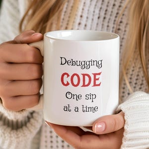 Może przedstawiać: Biały kubek ceramiczny z czarnym tekstem, kt&oacute;ry brzmi "Debugging CODE One sip at a time". Słowo "CODE" jest czerwone.