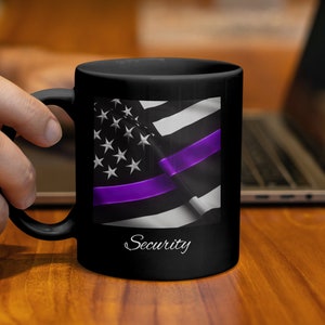 May include: Black coffee mug featuring a design of the American flag with a purple stripe. The mug has the word "Security" in a script font. The mug is held by a person.