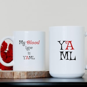 May include: Two white ceramic mugs with black and red text. The mug on the left says "My Blood Type is YAML". The mug on the right says "YAML" with the "A" in red.