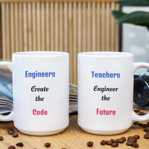 May include: Two white ceramic mugs with black handles. One mug reads "Engineers Create the Code" in blue, black, and red text. The other mug reads "Teachers Engineer the Future" in blue, black, and red text. Coffee beans are scattered around the mugs.