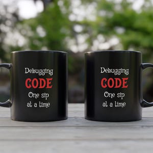 Może przedstawiać: Dwa czarne kubki do kawy z białym tekstem, kt&oacute;ry brzmi "Debugging CODE One sip at a time".