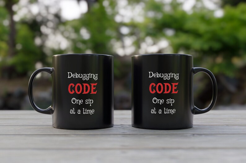 Może przedstawiać: Dwa czarne kubki do kawy z białym tekstem, kt&oacute;ry brzmi "Debugging CODE One sip at a time".