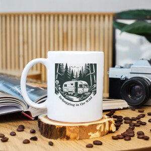 May include: White ceramic mug featuring a black and white illustration of a vintage camper in a forest scene. The mug has the text "Debugging in the Wild". The mug is on a wooden coaster, with coffee beans scattered around.