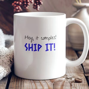 May include: White ceramic coffee mug with a blue inscription that reads "Hey, it compiles! SHIP IT!"