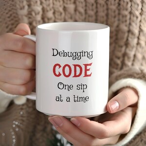 Może przedstawiać: Biały kubek ceramiczny z czarnym tekstem, kt&oacute;ry brzmi "Debugging CODE One sip at a time". Słowo "CODE" jest czerwone.