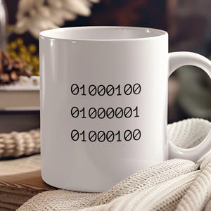 May include: White ceramic mug with black text that reads "01000100 01000001 01000100". The mug is sitting on a white knitted blanket.