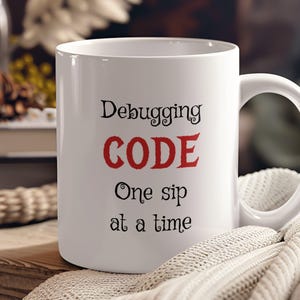 Może przedstawiać: Biały ceramiczny kubek do kawy z napisem "Debugging CODE One sip at a time" w czarnym i czerwonym kolorze.
