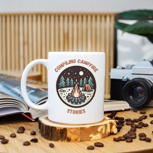 May include: White ceramic mug with a campfire scene and the text "Compiling Campfire Stories". The mug is sitting on a wooden coaster and there are coffee beans scattered around it.