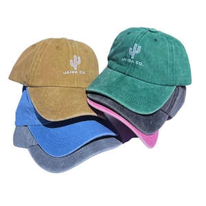 Pode incluir: Um conjunto de quatro bonés de beisebol de diferentes cores. O boné superior é verde com um cacto branco e o texto "JAIGA CO." bordado na frente. O segundo boné é castanho, o terceiro é azul e o boné inferior é preto.