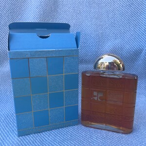 May include: A brown glass bottle of cologne with a silver cap. The bottle has a grid pattern on the front and the letter "C" in a square on the front. The bottle is sitting on a blue surface.
