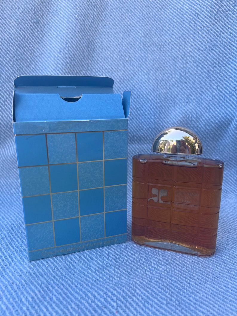 May include: A brown glass bottle of cologne with a silver cap. The bottle has a grid pattern on the front and the letter "C" in a square on the front. The bottle is sitting on a blue surface.
