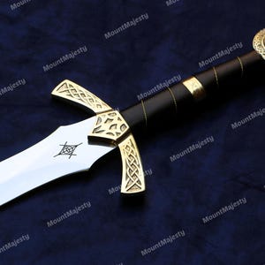 May include: A gold and silver sword with a black leather handle and a red jewel on the pommel. The sword has a Celtic knot design on the hilt and a silver blade.