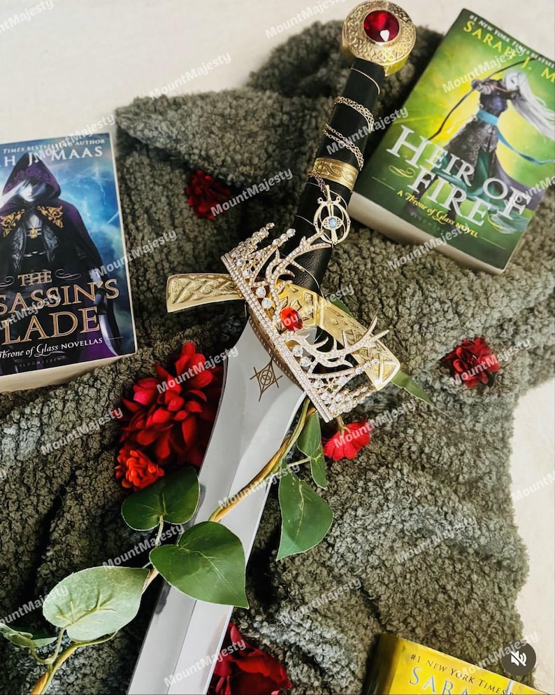 May include: A silver sword with a gold hilt and a red jewel on top. The sword is surrounded by a crown, red roses, and two books with the titles "The Assassin's Blade" and "Heir of Fire".