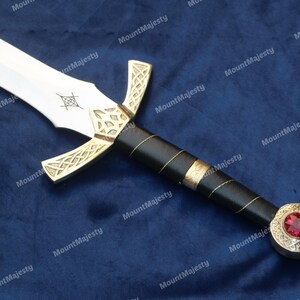May include: A silver sword with a black handle and a gold-coloured hilt. The hilt has a red jewel in the centre. The sword is on a blue fabric background.