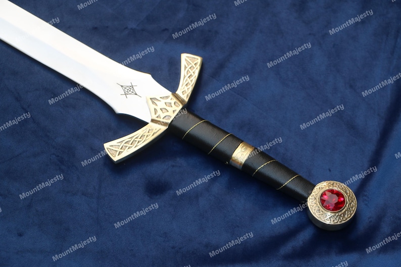 May include: A silver sword with a black handle and a gold-coloured hilt. The hilt has a red jewel in the centre. The sword is on a blue fabric background.