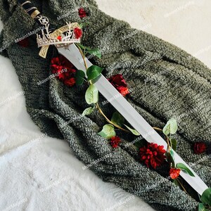 May include: A silver sword with a black handle and a gold crown on top, lying on a green fuzzy blanket with red roses.