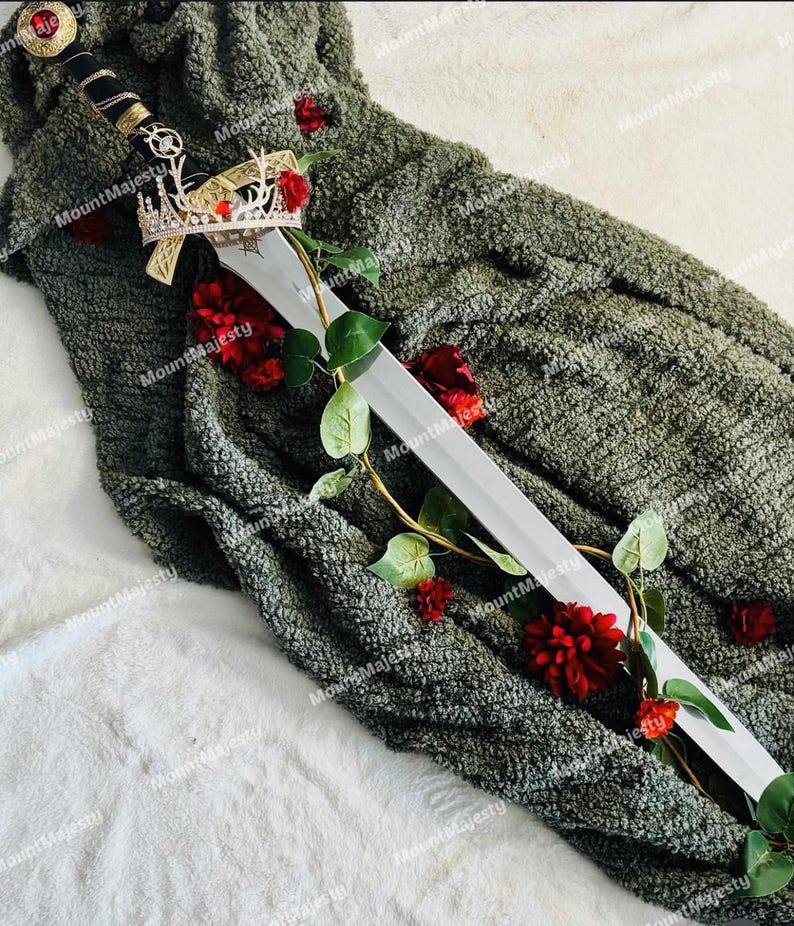 May include: A silver sword with a black handle and a gold crown on top, lying on a green fuzzy blanket with red roses.