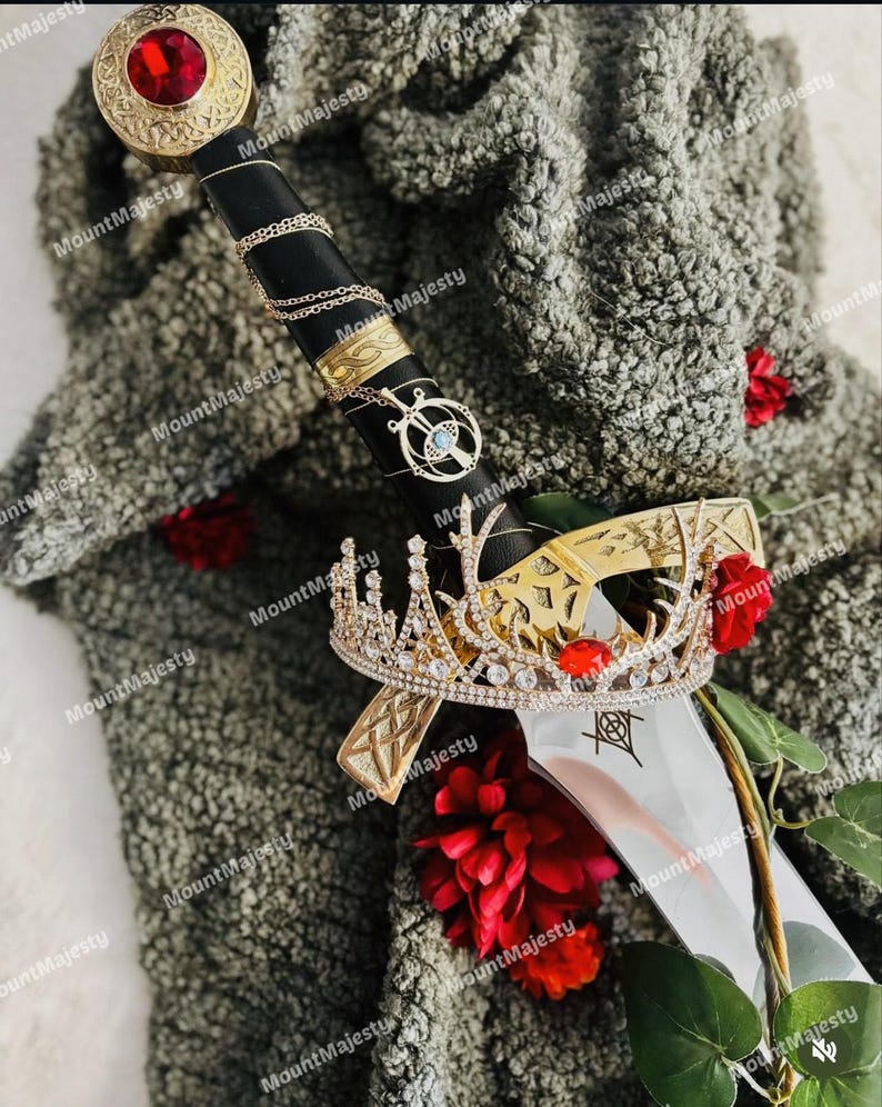 May include: A silver sword with a black handle and a gold crown on top. The sword is surrounded by red roses and a gray fuzzy fabric.