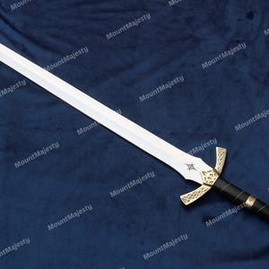 May include: A silver sword with a gold hilt and a red jewel on the pommel. The sword is lying on a blue fabric background.