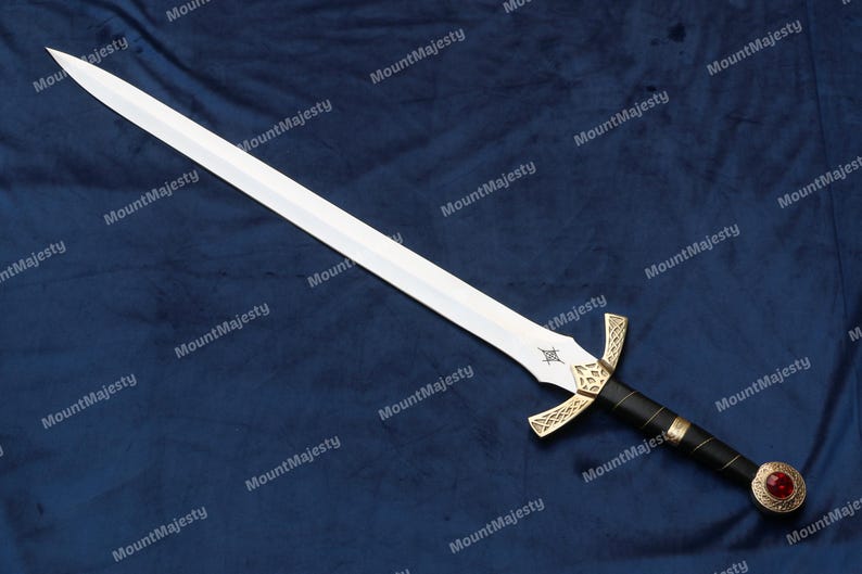 May include: A silver sword with a gold hilt and a red jewel on the pommel. The sword is lying on a blue fabric background.