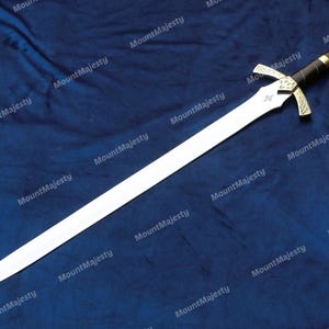 May include: A silver sword with a gold hilt and a red jewel on the pommel. The sword is lying on a blue fabric background.